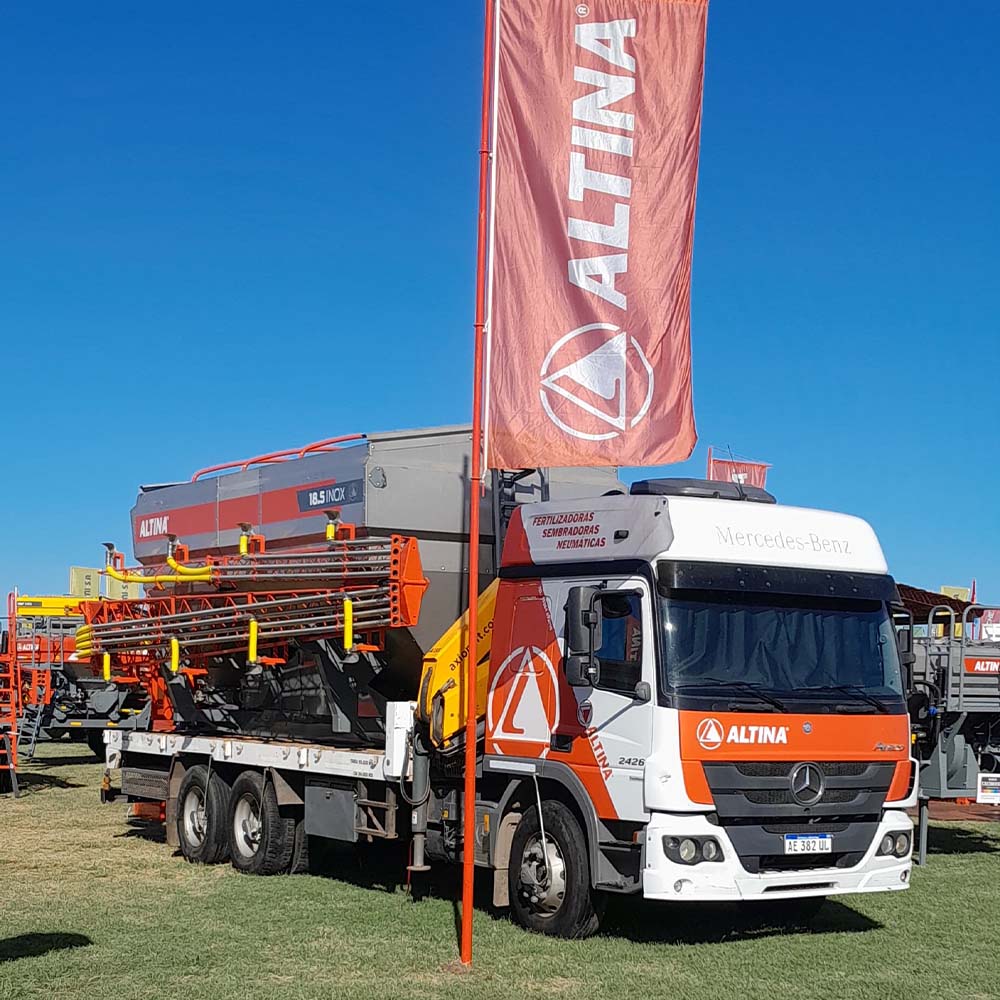 Camión Mercedes-Benz con gráfica de Altina transportando un kit Altina 18.5 INOX montado sobre la plataforma. El equipo agrícola, de gran porte, está diseñado para fertilización y siembra neumática. La imagen fue tomada en una exposición a campo, con bandera de la marca ondeando en primer plano.